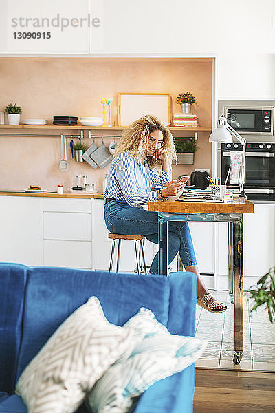 Eine Frau in voller Länge benutzt ein Smartphone  während sie in der Küche am Tisch sitzt