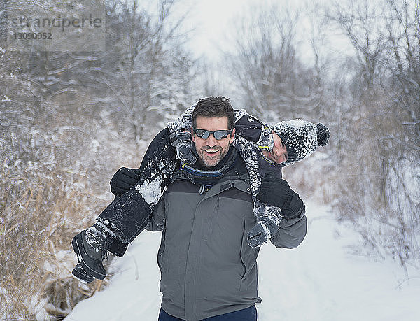 Spielerischer Vater trägt Sohn auf den Schultern auf verschneitem Feld gegen kahle Bäume