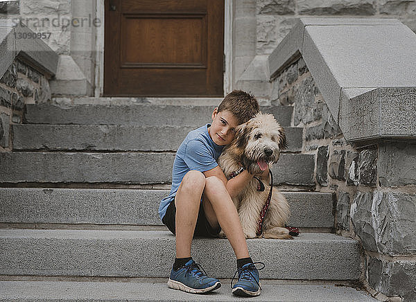 Porträt eines Jungen  der einen Hund umarmt  während er auf einer Treppe sitzt