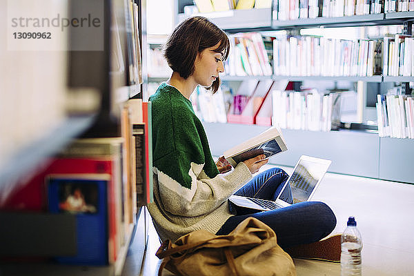 Seitenansicht einer Frau mit Laptop-Computer  die in der Bibliothek auf dem Boden sitzend ein Buch liest