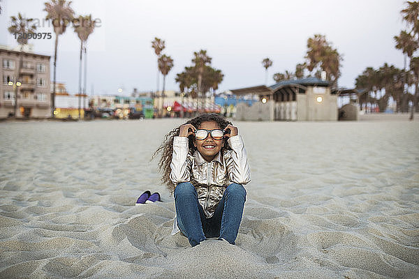 Glückliches Mädchen mit Sonnenbrille  das am Strand im Sand sitzt