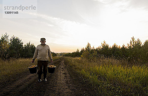Mann hält Pilze in Eimern  während er auf Feldweg auf Feld gegen Himmel steht