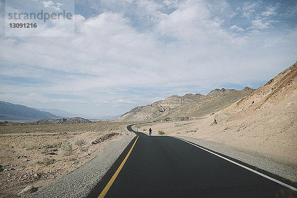 Fernansicht von Vater und Sohn beim Fahrradfahren auf einer Wüstenstraße vor bewölktem Himmel