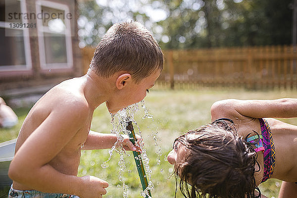 Verspielte Schwester schaut dem Bruder beim Bücken im Hof zu  wie er durch den Gartenschlauch Wasser trinkt