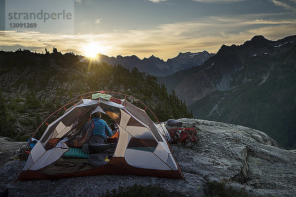 Frau sitzt bei Sonnenuntergang im Zelt auf einer Klippe vor Bergen