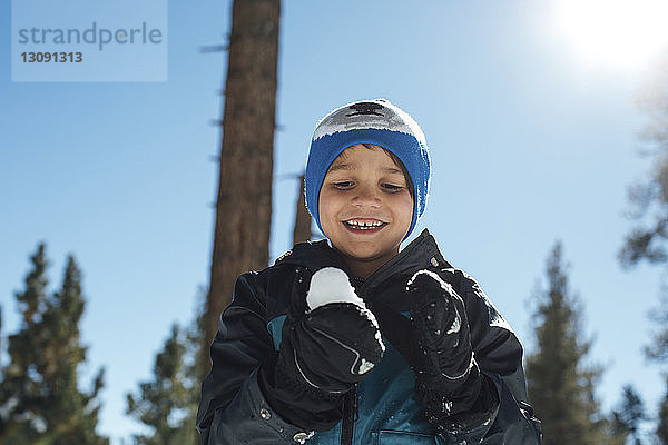 Tiefwinkelansicht eines glücklichen Jungen  der mit Schnee gegen den blauen Himmel im Wald spielt