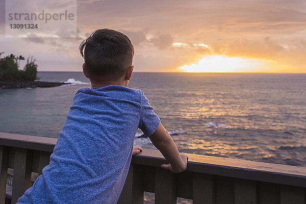 Rückansicht eines Jungen  der bei Sonnenuntergang an der Reling am Pier steht und aufs Meer schaut