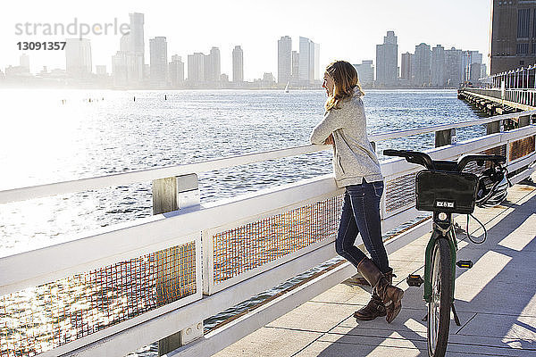 Nachdenkliche Frau mit Fahrrad steht auf Brücke am Fluss in der Stadt