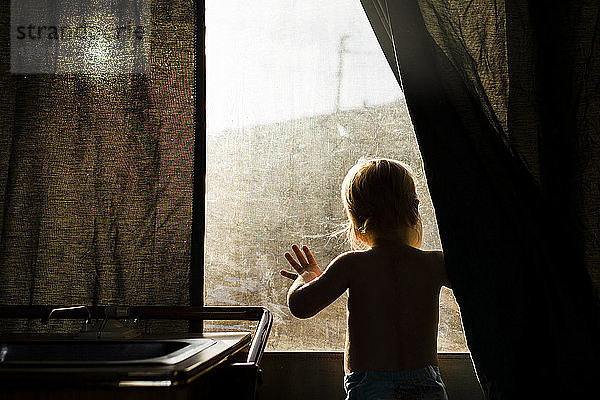 Rückansicht eines zu Hause stehenden Jungen  der durch ein Fenster schaut