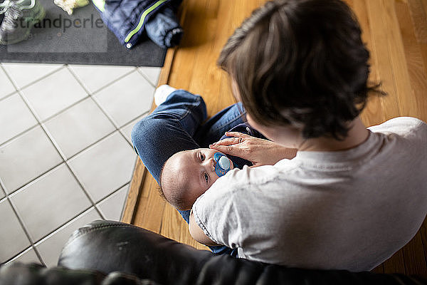 Hochwinkelaufnahme einer Mutter  die ihren Sohn hält  während sie zu Hause auf einem Parkettboden sitzt