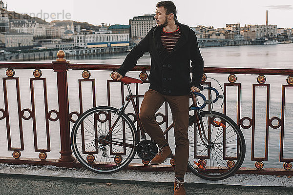 Radfahrer schaut weg  während er bei Sonnenuntergang mit dem Fahrrad auf der Brücke über den Fluss in der Stadt steht