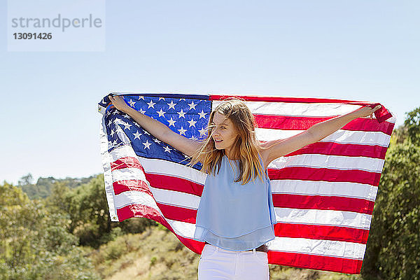 Glückliche Frau mit amerikanischer Flagge steht auf dem Feld vor klarem Himmel