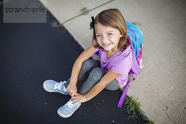 Hochwinkelporträt eines lächelnden Mädchens mit Rucksack  das auf einem Fußweg sitzt