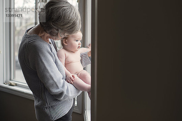 Großmutter hält hemdlose Enkelin  während sie zu Hause am Fenster steht