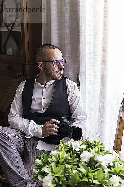 Nachdenklicher männlicher Fotograf mit Kamera sitzt zu Hause am Fenster