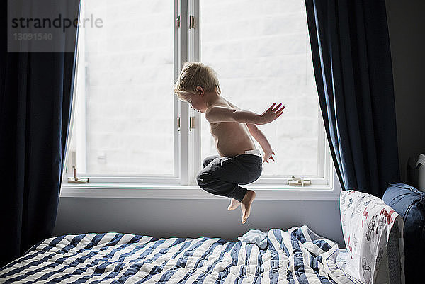 Junge ohne Hemd  der zu Hause am Fenster aufs Bett springt