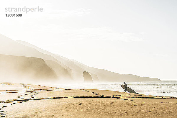 Mann mit Surfbrett steht am Strand am Ufer vor klarem Himmel