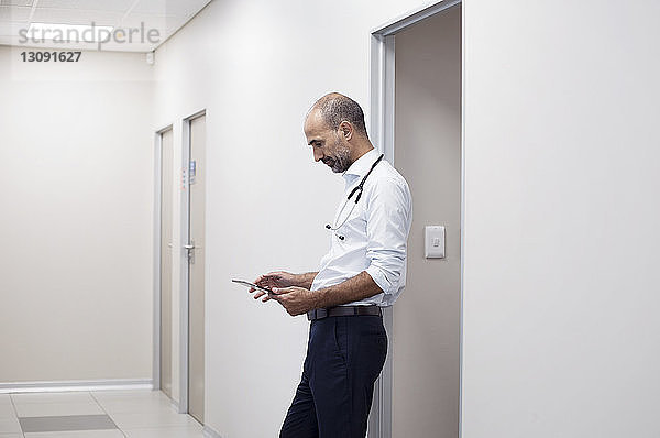 Seitenansicht eines Arztes  der einen Tablet-Computer benutzt  während er an der Tür im Korridor steht