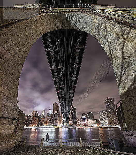 Mann sitzt auf Geländer unter Brücke am Fluss und beleuchtete Stadt in der Abenddämmerung
