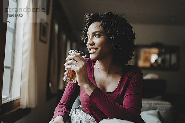 Nachdenkliche Frau trinkt  während sie zu Hause durchs Fenster schaut