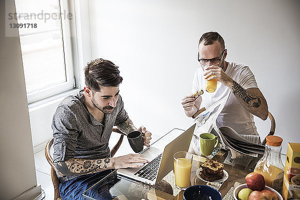 Schwule Männer benutzen Laptop und lesen Zeitung  während sie zu Hause frühstücken