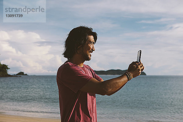 Seitenansicht eines Mannes  der mit einem Mobiltelefon am Strand vor bewölktem Himmel fotografiert