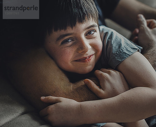 Mittelteil eines Vaters  der seinen glücklichen Sohn umarmt  während er zu Hause auf dem Sofa liegt