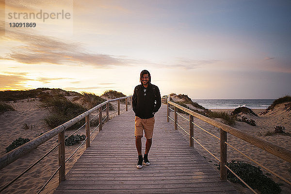 Lächelnder Wanderer  der bei Sonnenuntergang auf einem Steg am Strand gegen den Himmel läuft