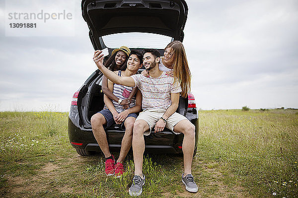 Fröhliche Freunde  die sich im Kofferraum eines Autos auf dem Feld gegen den Himmel im Smartphone bewegen