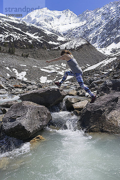 Seitenansicht einer Frau  die auf Felsen am Fluss gegen schneebedeckte Berge springt