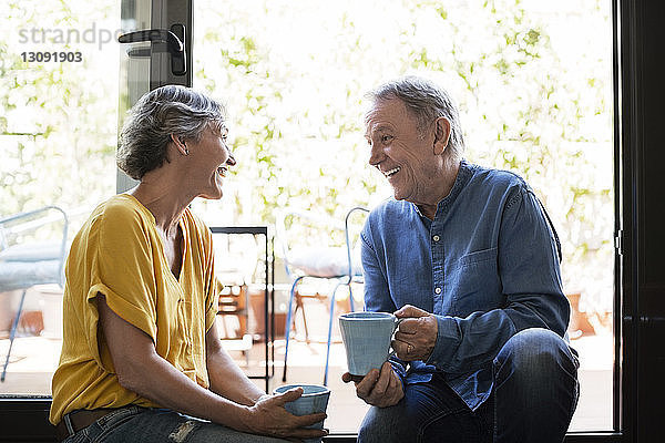 Fröhliches älteres Ehepaar unterhält sich  während es zu Hause Kaffeetassen am Fenster hält