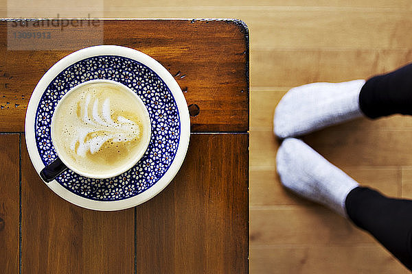 Draufsicht auf einen Cappuccino  der auf einem Holztisch von Frauenbeinen serviert wird