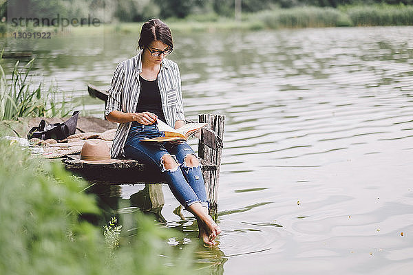 Frau liest Buch  während sie auf dem Pier über dem See im Wald sitzt