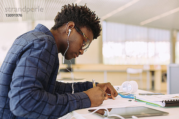 Mann mit Kopfhörern schreibt  während er am Tisch in der Bibliothek sitzt