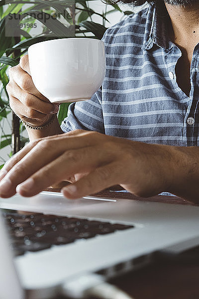 Mittendrin ein Mann  der eine Kaffeetasse hält  während er zu Hause einen Laptop auf dem Tisch benutzt