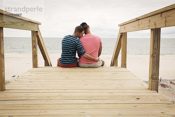 Rückansicht eines homosexuellen Paares  das auf einer Strandpromenade gegen das Meer sitzt