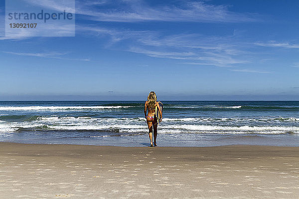 Rückansicht einer Frau im Bikini  die ein Surfbrett trägt  während sie am sonnigen Tag gegen den blauen Himmel in Richtung Meer läuft
