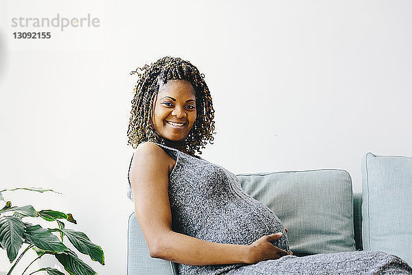 Porträt einer lächelnden schwangeren Frau  die zu Hause auf dem Sofa an der Wand sitzt