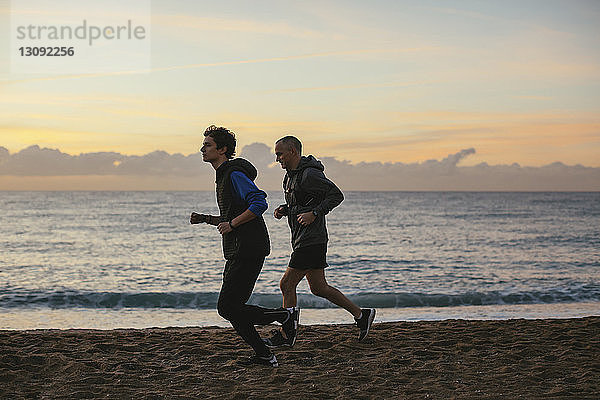 Seitenansicht von Vater und Sohn  die bei Sonnenuntergang am Strand gegen den Himmel joggen