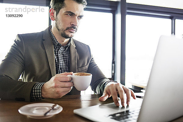 Konzentrierter Geschäftsmann hält Kaffeetasse  während er im Café am Laptop arbeitet
