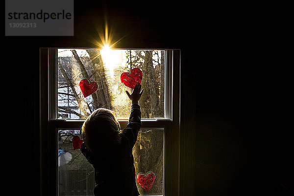 Rückansicht eines Jungen  der zu Hause Herzformen an ein Fenster klebt