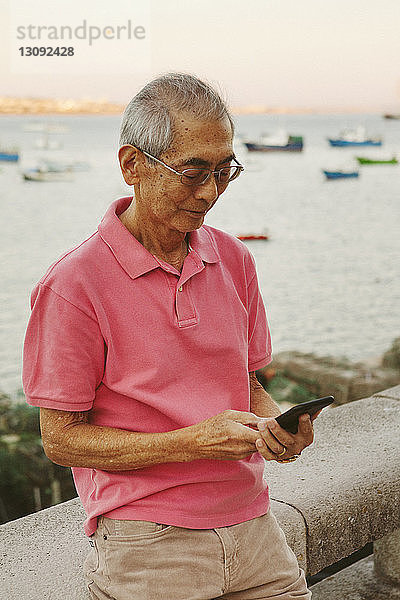 Älterer Mann benutzt Smartphone  während er sich an der Reling gegen das Meer lehnt