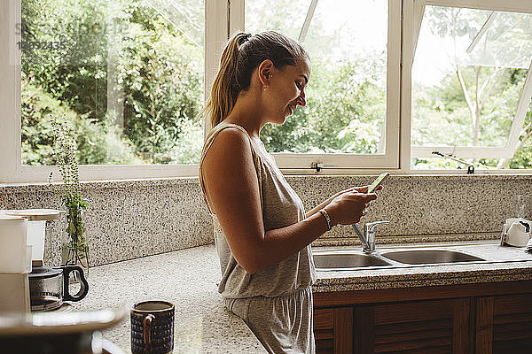 Seitenansicht einer lächelnden Frau  die ein Smartphone benutzt  während sie in der Küche steht