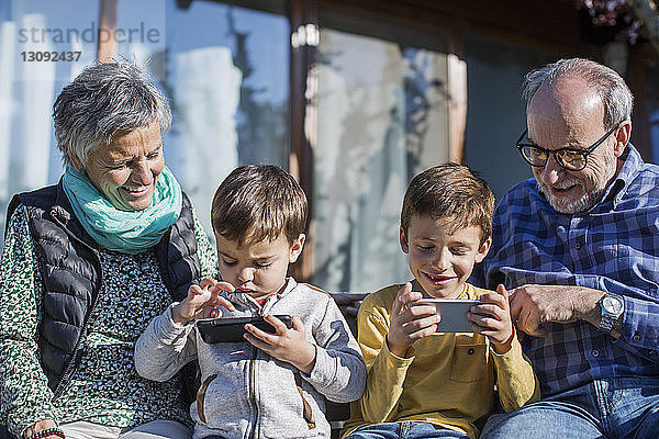 Glückliche Großeltern sehen Enkel an  die im Park Smartphones benutzen