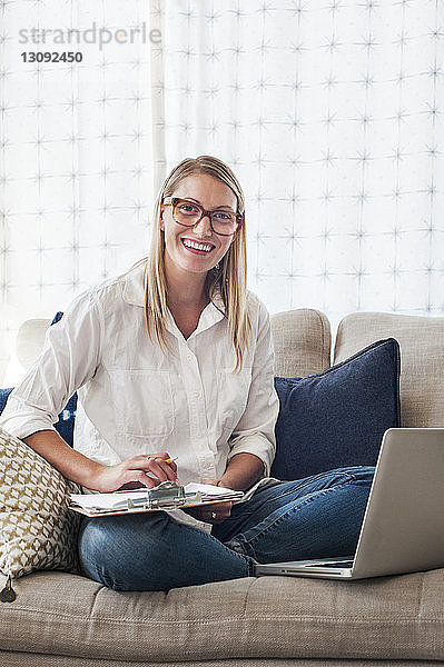 Porträt einer glücklichen Frau  die das Klemmbrett hält  während sie zu Hause auf dem Sofa sitzt