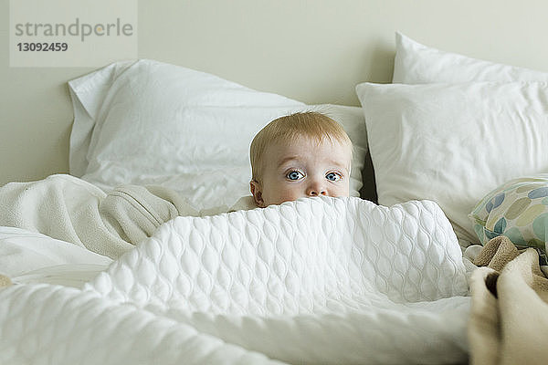 Süßer kleiner Junge versteckt sich zu Hause inmitten der Bettdecke auf dem Bett