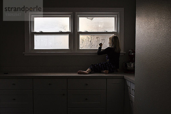 Mädchen macht Herzform am verdichteten Fenster  während sie zu Hause auf der Küchentheke sitzt