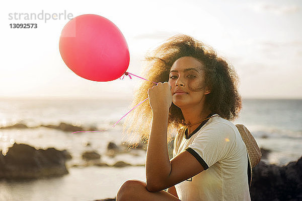 Porträt einer Teenagerin  die einen Ballon hält  während sie am Ufer gegen den Himmel sitzt