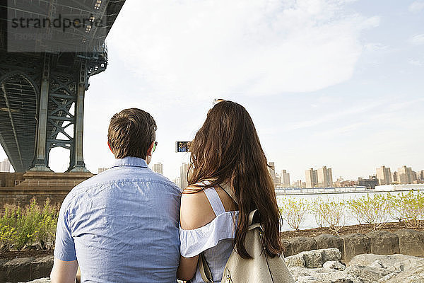 Rückansicht eines Paares  das telefoniert  während es unter der Brücke von Manhattan gegen den Himmel sitzt