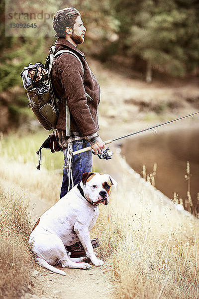 Seitenansicht eines Reisenden beim Angeln  während er mit seinem Hund am Seeufer sitzt